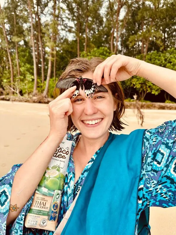 Meda Andreea Căpîlna râzând, cu un fluture așezat pe frunte, pe o plajă mărginită de vegetație